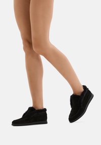 Calzature slip-on in suede nero con un bordo esterno morbido e peloso. Presentano una punta arrotondata e una suola piatta in gomma, progettate per il comfort.