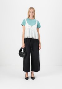 Teal t-shirt gelaagd met een wit topje met kant, gecombineerd met zwarte wijde broek, terwijl ze een kleine zwarte tas vasthoudt en puntige hakken draagt.