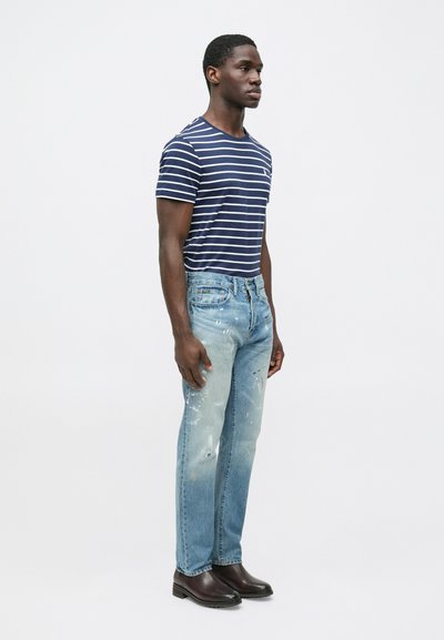 Prugasta mornarsko plava majica, svijetloplave poderane traperice s mrljama od boje i smeđe gležnjače. Model stoji na neutralnoj pozadini.