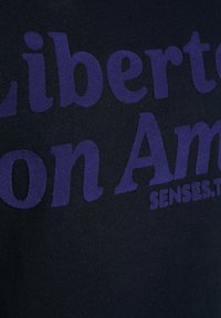 Schwarzes Kurzarmhemd mit fettem lila Text, der "Liberté on Amour" liest, mit leicht strukturiertem Material. Das Design umfasst eine abgerundete, moderne Schriftart.