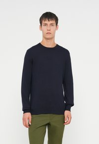 Suéter de malha azul-marinho com gola redonda e mangas longas. Punhos e barra canelados, confeccionado em tecido de malha lisa. Combinado com calças verde-azeitona.