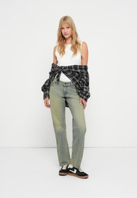 Top branco sem mangas, jeans soltos verde-claro e uma camisa xadrez preta atada à cintura. Ténis pretos com sola castanha.