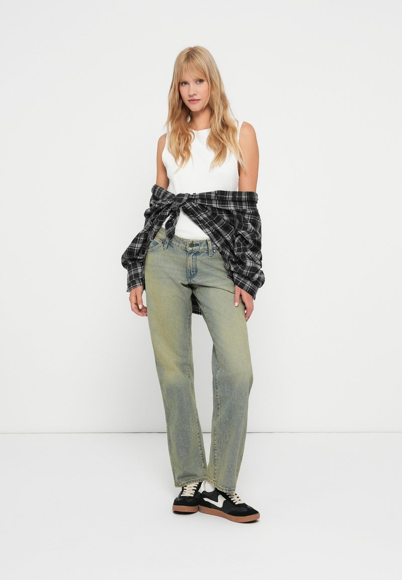 Top bianco senza maniche, jeans larghi verde chiaro e una camicia a quadri nera legata in vita. Sneakers nere con suola marrone.