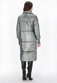 Lang, polstret frakk i metallic grå med høj krave. Har vandret syninger og ribbede manchetter. Sammen med knæhøje sorte støvler.