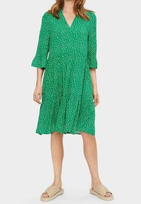 Zelené šaty po kolena s V-výstřihem, 3/4 rukávy a bílými puntíky. Rozšířený lem a lehký materiál. Nosené s béžovými šlapkami.