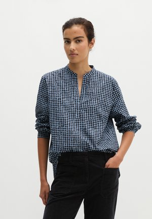 Frau mit dunklem, zurückgebundenem Haar trägt eine blau-schwarze gepunktete Bluse und schwarze Hose, steht mit einer Hand in der Tasche vor weißem Hintergrund.