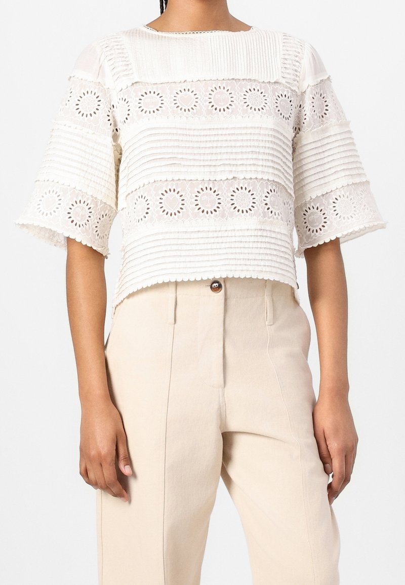 Femme portant un haut blanc à manches courtes avec une dentelle florale à œillets et un tissu plissé, associé à un pantalon beige taille haute.