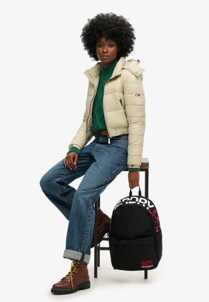 Une jeune femme aux cheveux afro porte une doudoune crème, un pull vert, un jean retroussé et des bottes marron, assise sur un tabouret en tenant un sac à dos Superdry noir.