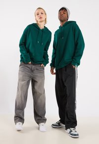 To personer iført grønne hettegensere, den ene med lommer foran, og stilige grå baggy jeans, den andre i svarte joggebukser med striper og sneakers.