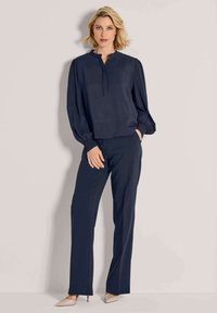 Frau, die gegen eine Wand steht, trägt eine marineblaue Bluse mit puffigen Ärmeln und passende marineblaue Hosen, beige spitze Absatzschuhe.