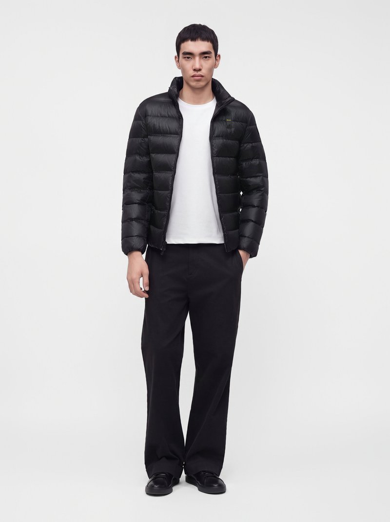 Jeune homme portant une doudoune noire, un t-shirt blanc, un pantalon noir et des chaussures noires, debout devant un fond clair uni.