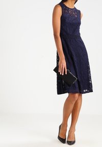 Navy lace dress with a fitted bodice and flared skirt, featuring a sheer lace neckline, paired with a black clutch and high heels.