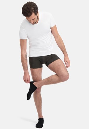 Schwarze Baumwoll-Boxershorts, weißes V-Ausschnitt-T-Shirt, schwarze Socken. Modell steht, ein Bein erhoben, mit sichtbaren Oberkörper-Tattoos. Einfaches Design.