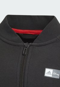 Giacca nera con zip e colletto alto, caratterizzata da un accento rosso all'interno e da una toppa con il logo bianco di Adidas e Disney sul petto.
