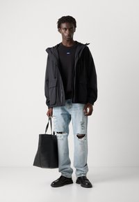 Black lightweight jacket with a hood, paired with a black sweater, distressed light blue jeans, and black shoes. Holding a black tote bag.