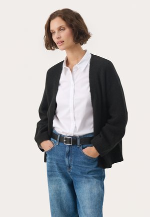 Schwarzer oversized Cardigan, weiche Textur, offenes Frontdesign, getragen über einem weißen Hemd mit Knöpfen und blauen Jeans, mit einem schwarzen Gürtel mit silberner Schnalle.
