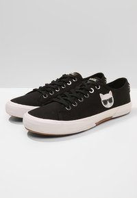 Mustat kangaskengät, joissa on valkoiset kumipohjat, mustat nauhat ja valkoinen kissa aurinkolasit päässä -kuva sivussa.