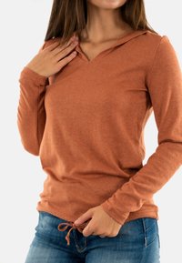 Donna che sistema il cordoncino dell'orlo del suo maglione a maniche lunghe color ruggine, indossando jeans blu, su uno sfondo semplice.