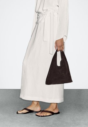 Person trägt einen weißen langen Rock und einen Pullover, hält eine dunkelbraune Wildlederhandtasche und trägt dunkelbraune flache Sandalen.