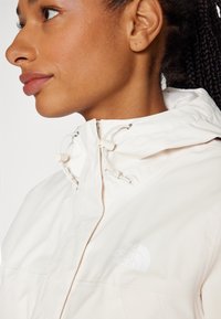 Giacca leggera di colore crema con cappuccio, dotata di chiusura con zip e coulisse. Include un sottile logo bianco sul petto.