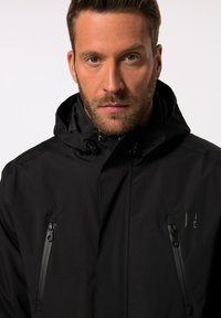 Sort vandtæt jakke med lynlåslukning foran, justerbar hætte og sidelommer med diskret branding. Glat overflade med mat finish.