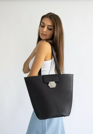Zwarte tote bag van soepel leer met een hexagonaal zilveren sluiting en platte schouderbanden, met een minimalistisch ontwerp en rechtopstaande vorm.