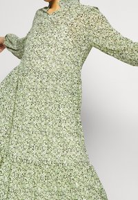 Pitkä vihreä kukkamekko, jossa on korkea kaulus, pitkät puffihihat ja levenevä helma. Kankaassa on läpikuultava rakenne, jossa on pieniä valkoisia ja vihreitä kuvioita.