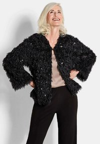 Schwarze Kunstfederjacke mit strukturiertem Flaum, im cropped Design und verstreuten Pailletten, kombiniert mit einem beigen Oberteil und schwarzen Hosen.