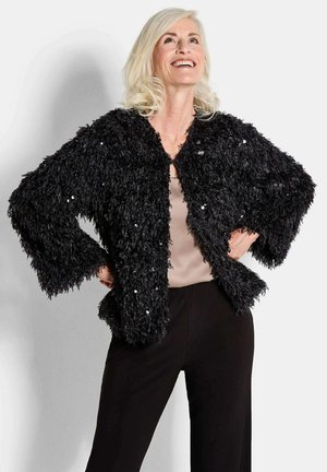 Zwarte nep bontjack met een textuur van pluizigheid, met een cropped ontwerp en verspreide pailletten, gecombineerd met een beige top en zwarte broek.