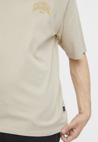 Beige bomull T-skjorte med korte ermer, rund halsutringning og "CASUAL"-tekst i gul brodering nær øvre venstre hjørne.