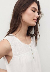 Blusa bianca senza maniche con un yoke ricamato testurizzato, scollo rotondo e bottoni decorativi sul davanti. Tessuto morbido e leggero.