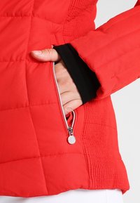 Chaqueta acolchada roja con un bolsillo lateral con cremallera, puños interiores negros y paneles texturizados en las mangas. Tela suave con detalles metálicos en la cremallera.