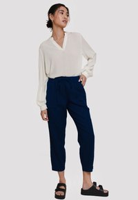 Witte blouse met lange mouwen en kraag, gecombineerd met donkerblauwe, losse broek met een trekkoord in de taille en voorzakken. Zwarte sandalen worden gedragen.