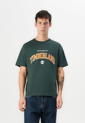 Dark green cotton t-shirt featuring a round neckline, short sleeves, and orange "TIMBERLAND" text with a logo above. Casual fit.