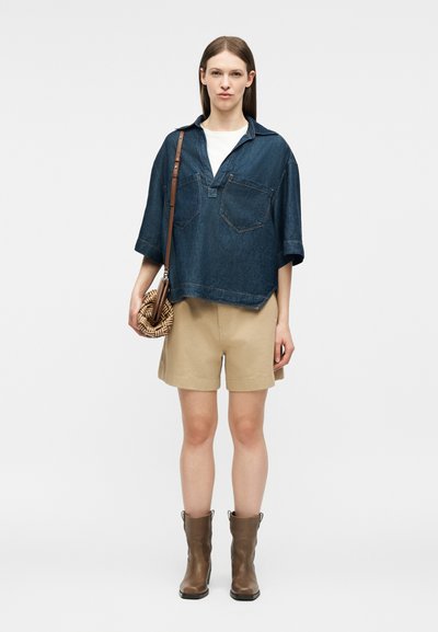 Γυναίκα που φοράει σκούρο τζιν πουκάμισο με κοντά μανίκια, μπεζ σορτς, καφέ μπότες αστραγάλου και κρατάει στρογγυλή πλεκτή τσάντα χιαστί.