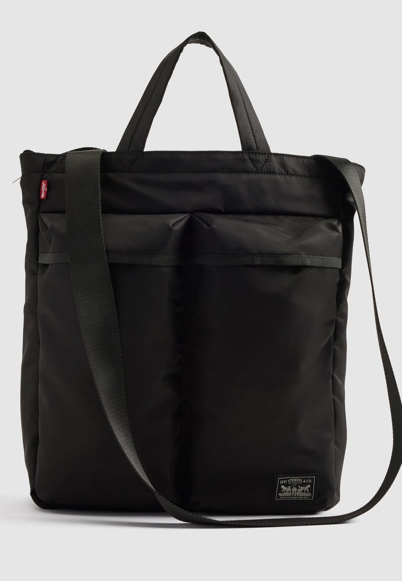 Bolsa tote negra de nylon con asas superiores dobles y una correa de hombro desmontable. Cuenta con bolsillos frontales y un parche con el logo en la base.