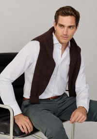 Wit geruite overhemden met een donkerbruine trui over de schouders, grijze broek en een bruine riem. Zittend op een zwarte stoel.