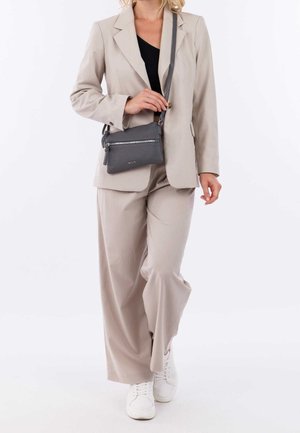 Kvinde i beige jakkesæt med sort top, der bærer en grå læder crossover-taske og har hvide sneakers på, går mod en hvid baggrund.