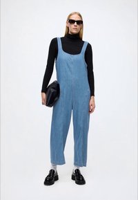 Vaaleansininen farkkuhaalari leveälahkeisella mallilla, säädettävillä hihnoilla ja kulmikkaalla pääntiellä, yhdistettynä mustaan poolopaitaan ja mustiin loafereihin.