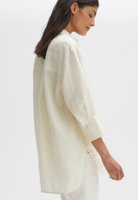 Light yellow linen button-up shirt with long sleeves, a relaxed fit, and subtle vertical texture. Features a curved hem and side slits.