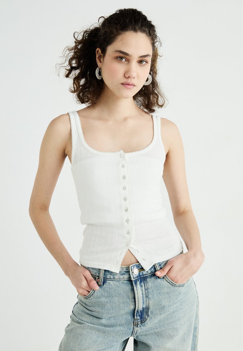 Top in bianco a coste con scollatura quadrata, caratterizzato da una chiusura frontale con bottoni e una delicata rifinitura. Abbinato a jeans larghi di colore blu chiaro.