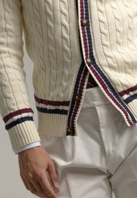 Cárdigan de punto de cable color crema con acentos a rayas en burdeos y azul marino, con botones de madera y puños acanalados, combinado con pantalones blancos.