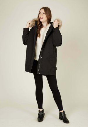 Parka noire avec une capuche en fourrure, dotée d'une fermeture éclair et de poches avant. Portée par-dessus un pull en tricot crème et un legging noir.