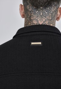 Chaqueta de tela negra con patrón de espiga y cuello estructurado. Presenta una etiqueta rectangular dorada de la marca en la parte posterior, cerca del escote.