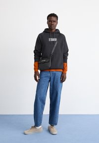 Fekete kapucnis pulóver logóval, narancssárga alsó réteg, kék farmer, szürke keresztpántos táska, bézs sportcipő, egyszínű háttér kék padlóval.