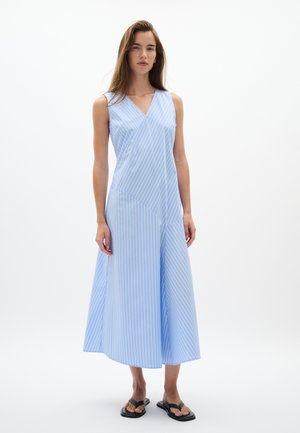 Mujer de pie con un vestido sin mangas azul claro con rayas verticales blancas y sandalias planas negras sobre un fondo blanco liso.