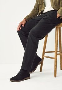 Chaussures en daim noir avec lacets, associées à un pantalon foncé, assis sur un tabouret en bois. Le design présente des lignes épurées et une texture lisse.