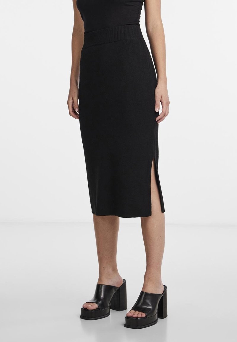 Femme portant une jupe midi noire avec une fente latérale et des sandales compensées noires à talons hauts, debout sur un fond blanc.