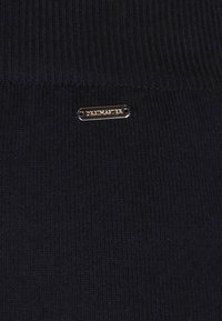 Dunkelblau gerippter Stoff mit einem kleinen goldenen Metalletikett, auf dem "DREIMASTER" eingraviert ist, was dem Textur und Design zusätzliche Details verleiht.