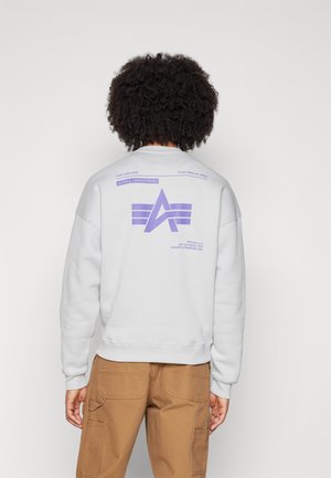 Person med krøllet hår iført hvid sweatshirt med lilla Alpha Industries-logo og tekst, kombineret med brune bukser, set bagfra.